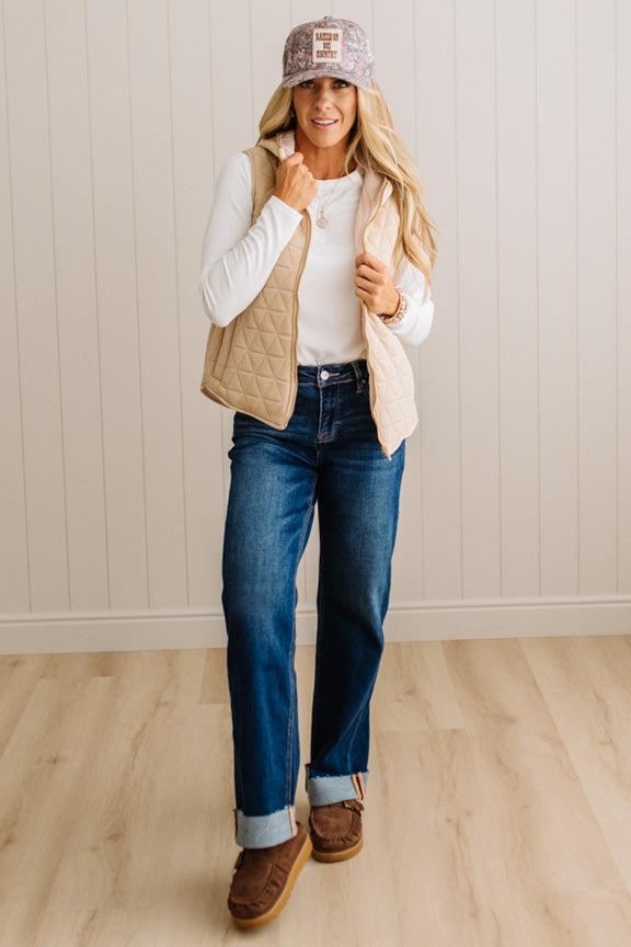 A woman modeling a beige quilted vest over a white long-sleeve top, paired with blue jeans and brown mule shoes, standing in a room with light wood floors and white paneled walls.