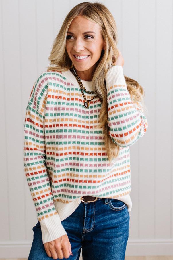 Blonde woman standing in brightly lit studio wearing a multi color sweater that has a relaxed, slightly cropped fit hits just right over high-waisted denim jeans.