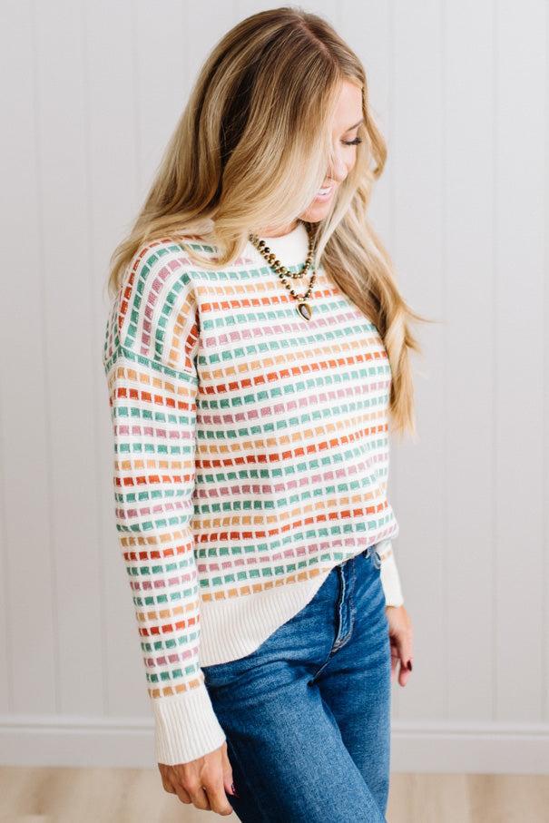 Blonde woman standing in brightly lit studio wearing a multi color sweater that has a relaxed, slightly cropped fit hits just right over high-waisted denim jeans.