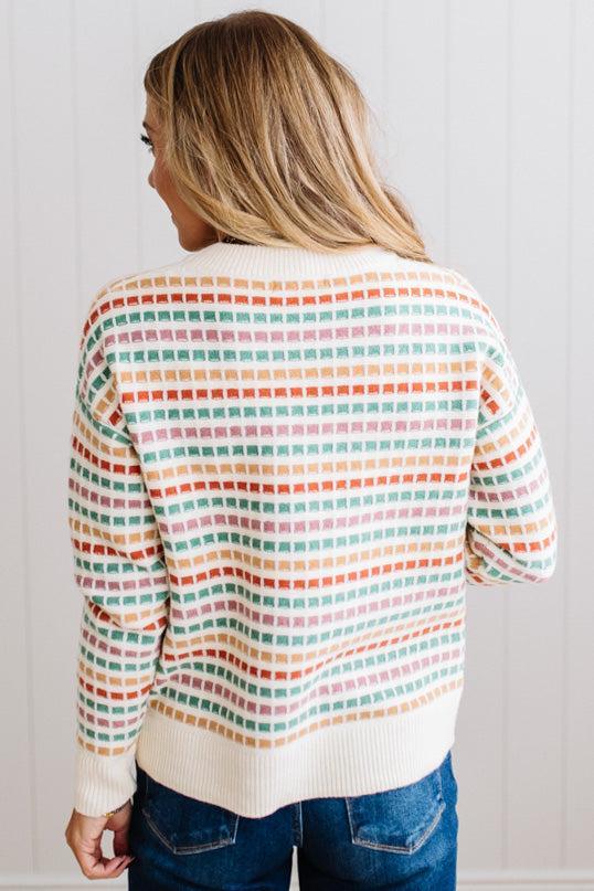 Blonde woman standing in brightly lit studio wearing a multi color sweater that has a relaxed, slightly cropped fit hits just right over high-waisted denim jeans.
