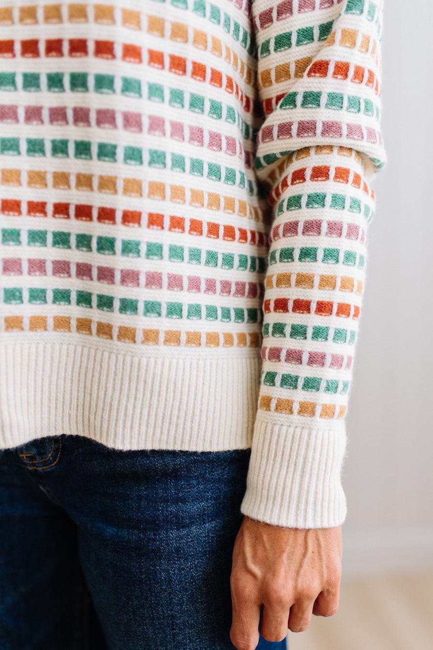 Blonde woman standing in brightly lit studio wearing a multi color sweater that has a relaxed, slightly cropped fit hits just right over high-waisted denim jeans.