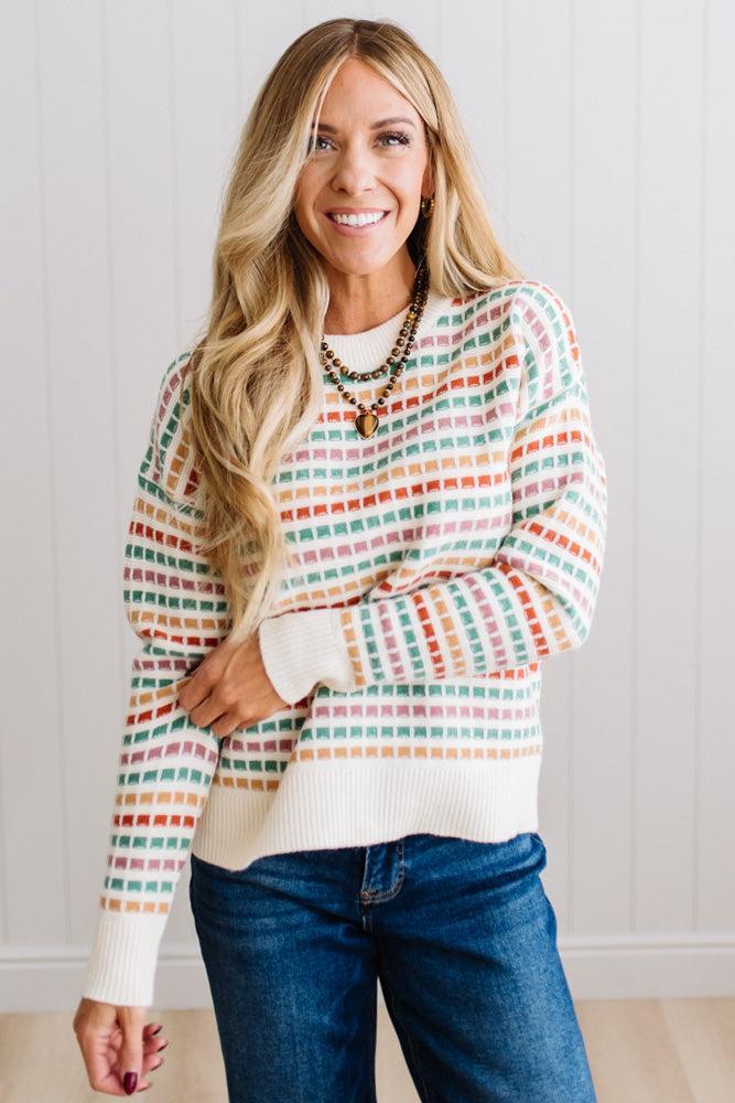 Blonde woman standing in brightly lit studio wearing a multi color sweater that has a relaxed, slightly cropped fit hits just right over high-waisted denim jeans.