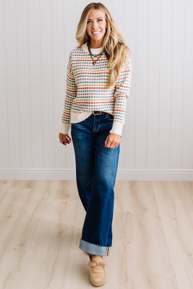 Blonde woman standing in brightly lit studio wearing a multi color sweater that has a relaxed, slightly cropped fit hits just right over high-waisted denim jeans.