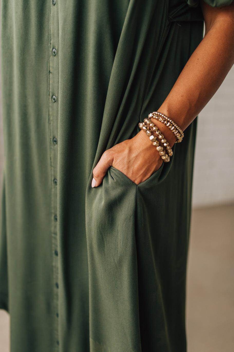 Person wearing a green dress with buttons and holding hands in pockets, wearing multiple bracelets.