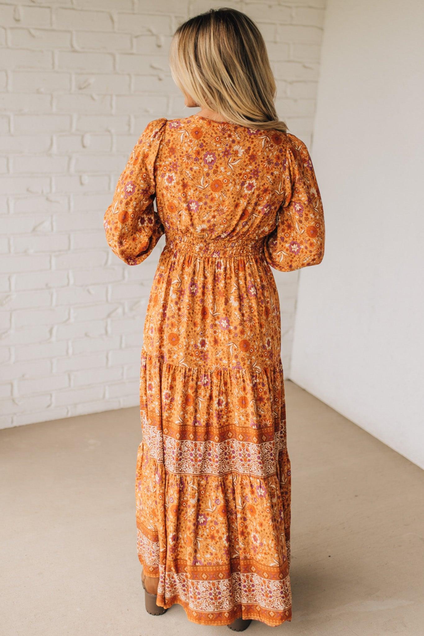 Woman wearing a long orange floral dress against a white brick wall.