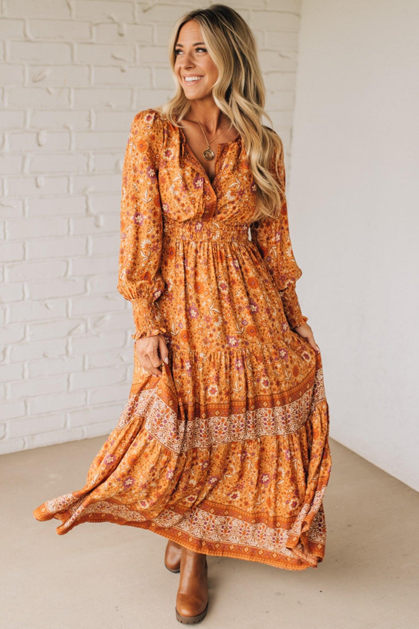 Woman wearing a long orange floral dress against a white brick wall.