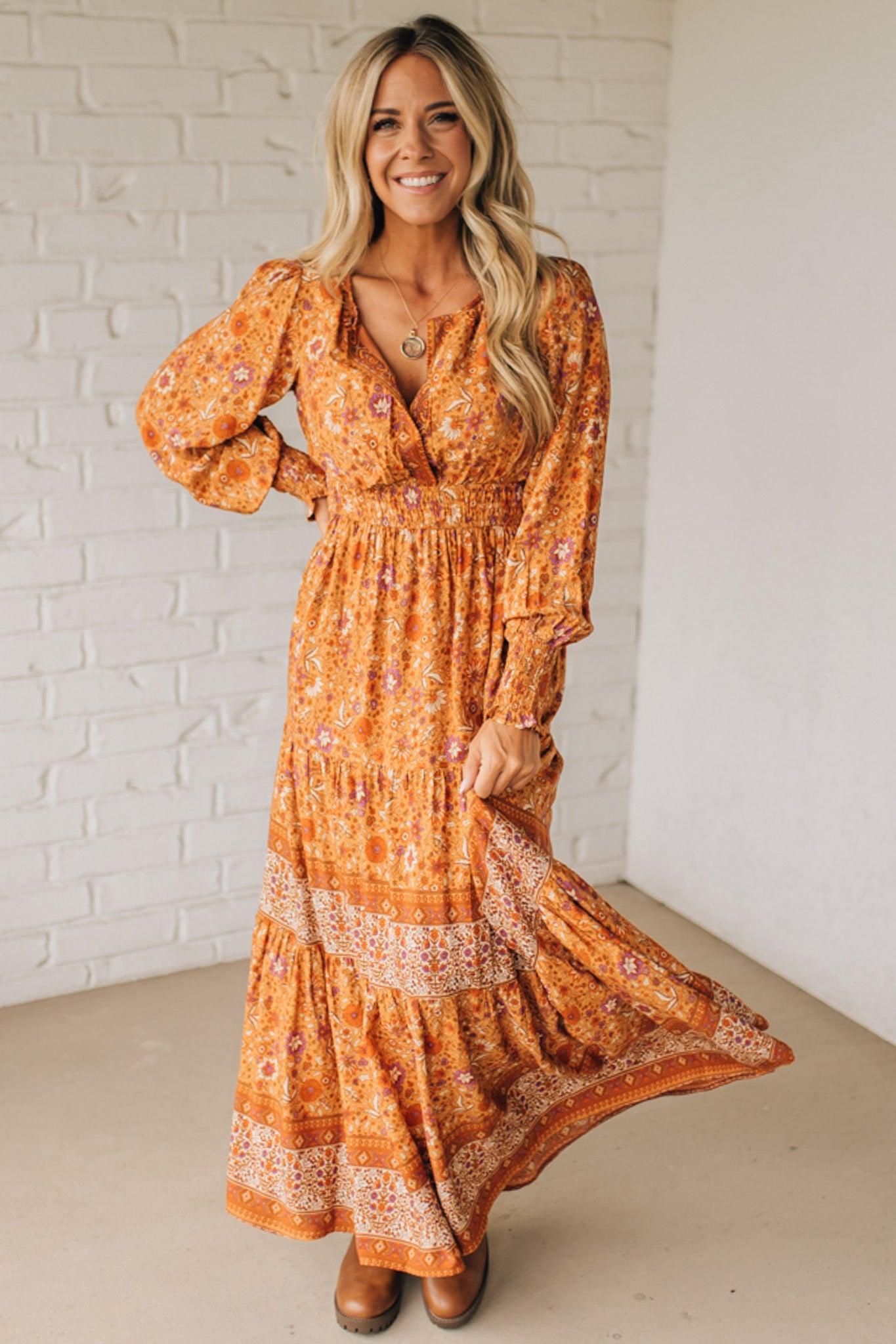 Woman wearing a long orange floral dress against a white brick wall.