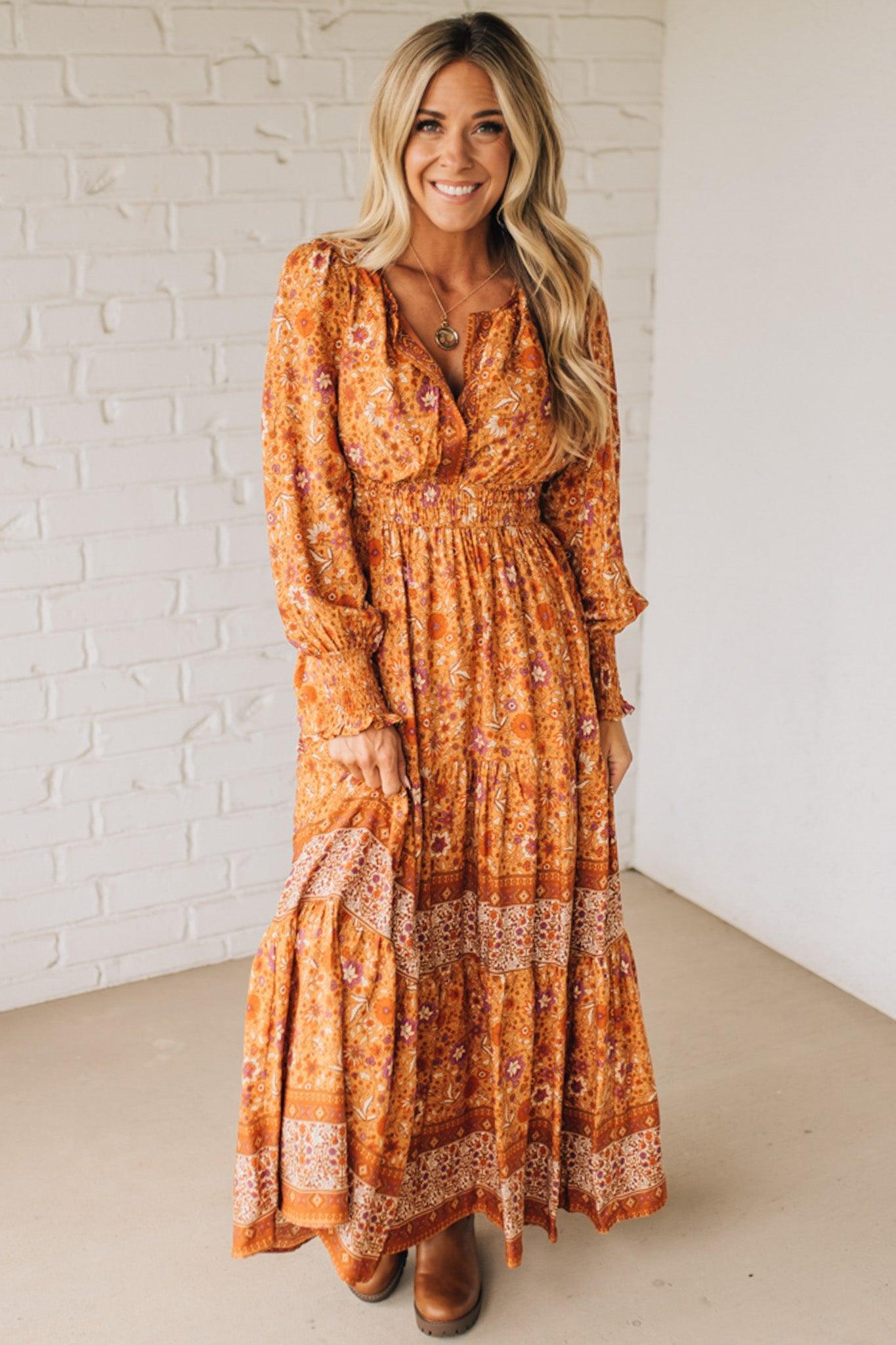 Woman wearing a long orange floral dress against a white brick wall.