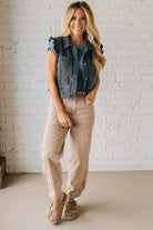 Woman wearing a denim vest, blue shirt, beige pants, and sandals against a white brick wall.