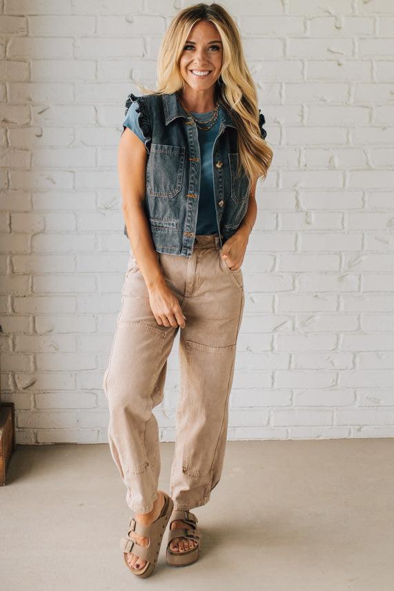 Woman wearing a denim vest, blue shirt, beige pants, and sandals against a white brick wall.