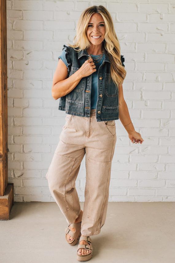 Woman wearing a denim vest over a blue shirt with beige pants against a white brick wall.