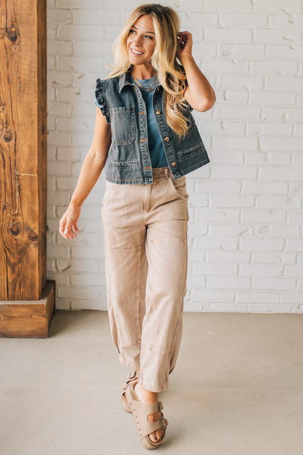 Woman wearing a denim vest and beige pants in a room with white brick walls and wooden furniture.