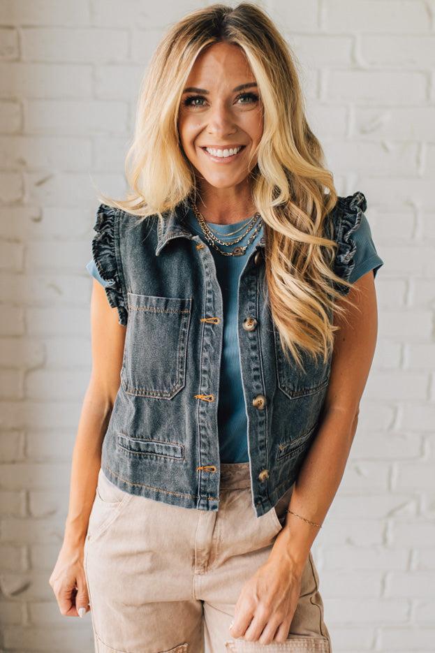 Woman wearing a denim vest over a blue top with beige pants against a white brick wall.