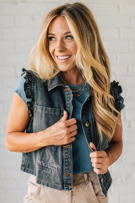Woman wearing a denim vest over a blue top against a white brick wall.