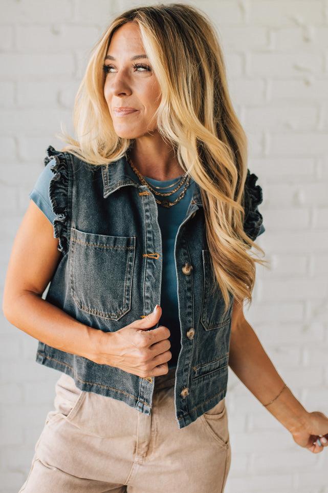 Woman wearing a denim vest over a blue top against a white brick wall.