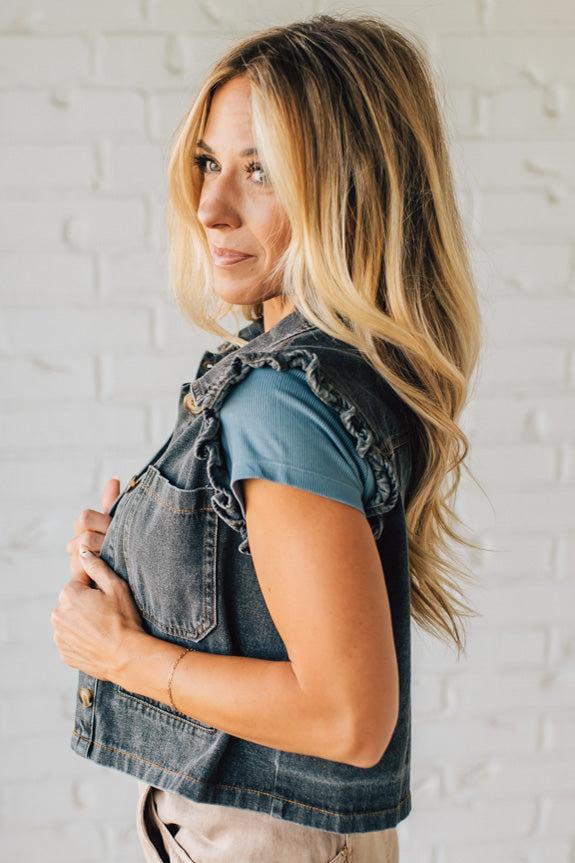 Woman wearing a denim vest over a teal top against a white brick wall.