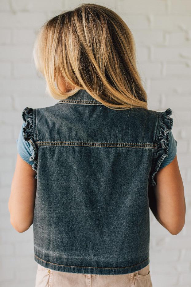 Person wearing a denim vest with ruffled shoulders against a light brick wall.
