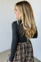 Woman wearing a charcoal top and plaid skirt against a white wall
