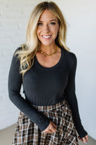 Woman wearing a charcoal long-sleeve top and plaid skirt against a white wall