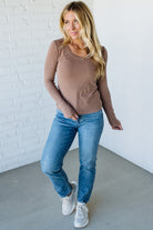 Woman wearing a brown top and blue jeans against a white brick wall.