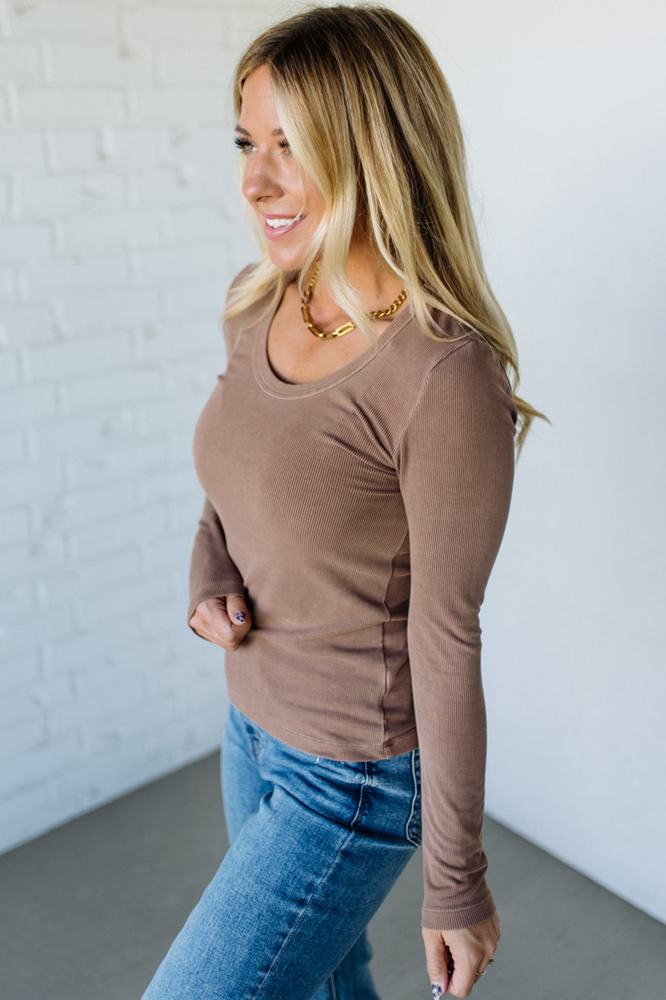 Woman wearing a brown long-sleeve top and blue jeans against a white brick wall.