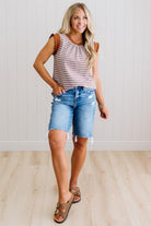 Woman wearing a striped tank top and denim shorts standing against a light wooden floor and white paneled wall.