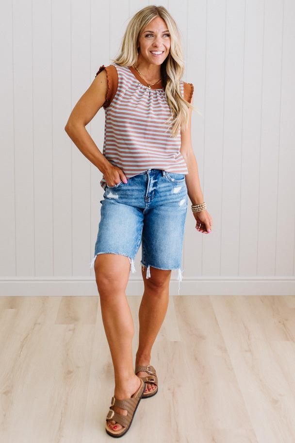 Woman wearing a striped tank top and denim shorts standing against a light wooden floor and white paneled wall.