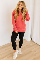 Woman wearing a coral sweatshirt and black leggings standing in a room with white walls and wooden floor.