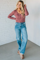 Woman wearing a pink long-sleeve top and blue jeans against a white brick wall.