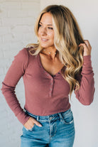 Woman wearing a mauve long-sleeve top and blue jeans against a white brick wall.