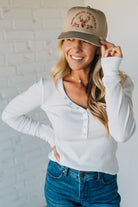 Woman wearing a beige cap with a logo and a white long-sleeve shirt against a white brick wall.