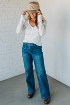 Woman wearing a white long-sleeve top, blue jeans, and a beige cap against a white brick wall.