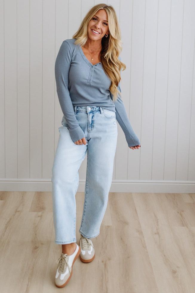 Woman wearing a blue top and light blue jeans standing against a white paneled wall.