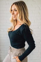 Woman wearing a black long-sleeve top and beige pants against a white brick wall.