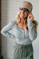 Woman wearing a beige cap and gray long-sleeve shirt against a white brick wall.