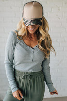Woman wearing a gray long-sleeve top and green pants with a beige and camouflage visor against a white brick wall.