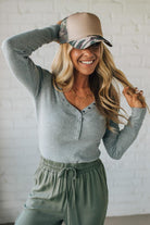 Woman wearing a gray long-sleeve top and green pants, smiling and adjusting a beige cap with a patterned visor against a white brick wall.