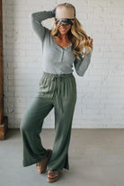 Woman wearing a gray long-sleeve top and green wide-leg pants against a white brick wall.
