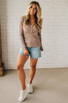 Woman wearing a beige long-sleeve top and denim shorts standing against a white brick wall.