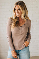 Woman wearing a brown long-sleeve top and blue jeans against a white brick wall.