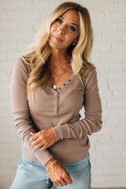 Woman wearing a beige long-sleeve top and blue jeans against a white brick wall.
