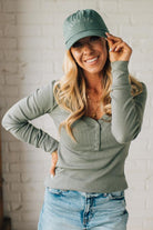 Woman wearing a green cap and long-sleeve shirt against a white brick wall.