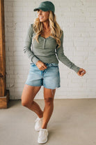 Woman wearing a green long-sleeve top, denim shorts, and white sneakers against a white brick wall.