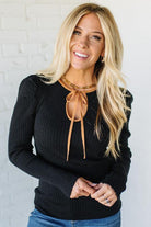 Woman wearing a black sweater with a brown tie neckline against a white brick wall.