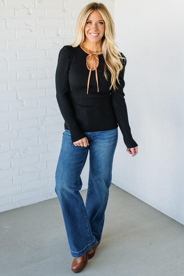 Woman wearing a black sweater with a brown bow and blue jeans against a white brick wall.