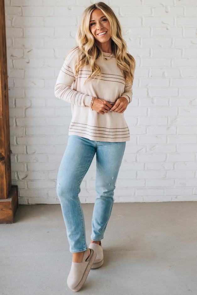 Woman wearing a striped sweater and jeans standing against a white brick wall.