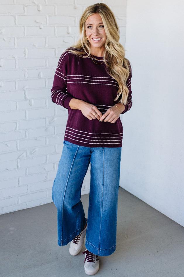 Woman wearing a maroon sweater with white stripes and blue jeans against a white brick wall.