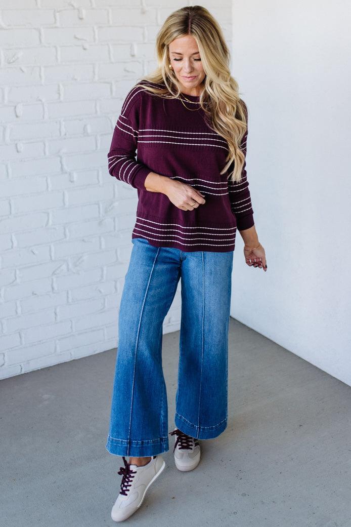 Woman wearing a burgundy sweater with white stripes and blue jeans against a white brick wall.