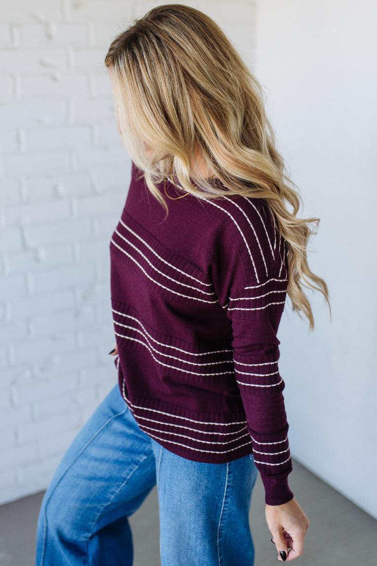 Person wearing a maroon sweater with white stripes and blue jeans against a white brick wall.