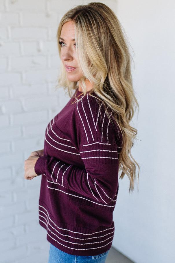 Woman wearing a maroon sweater with white stripes against a white brick wall.